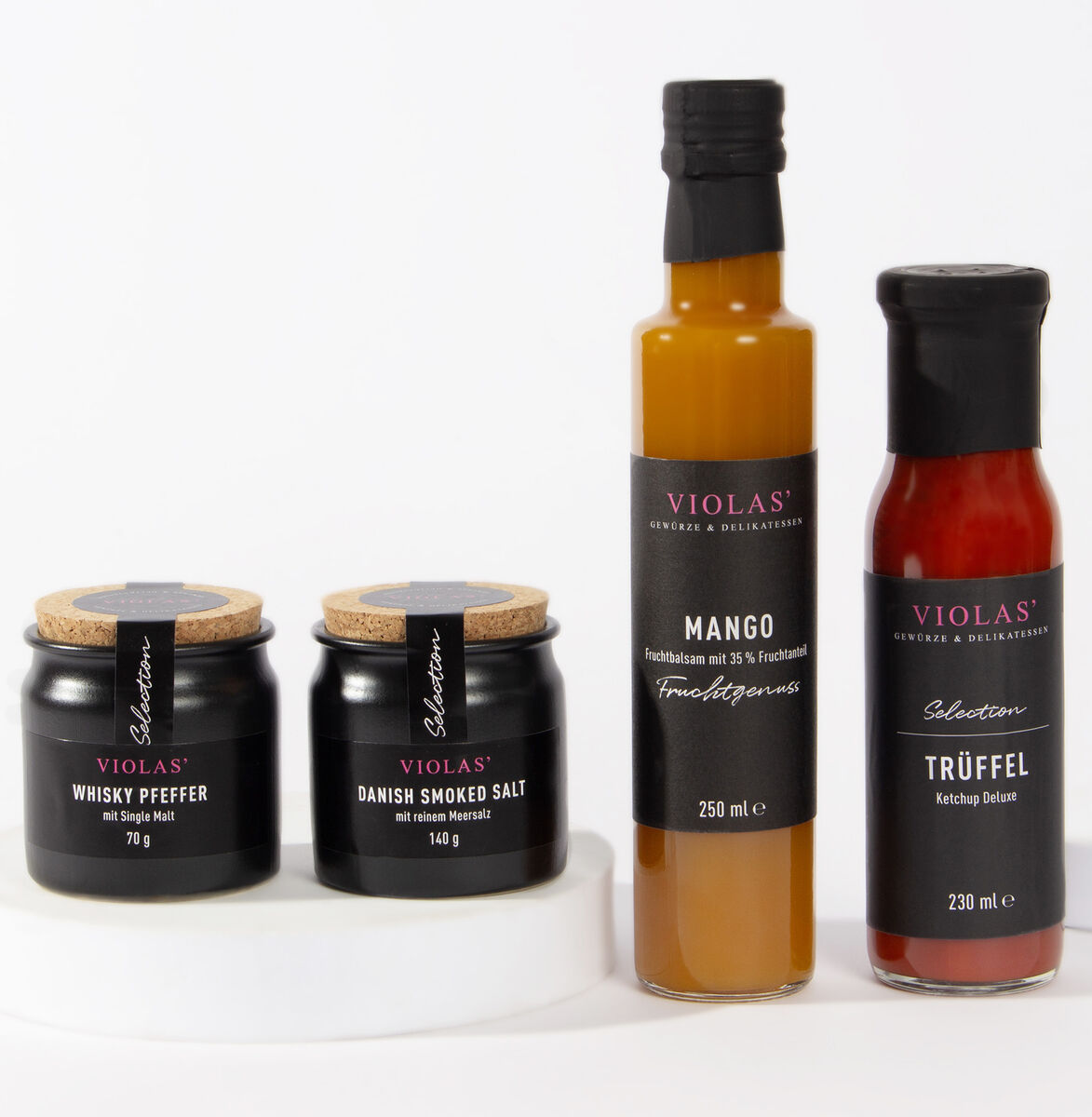 Foto eines ZEIT Genuss-Selection Sets mit vier verschiedenen Feinkostprodukten: zwei Gläser und zwei Flaschen. Von links nach rechts: Whisky Pfeffer, Dänisches Geräuchertes Salz, Mango Fruchtgenuss und Trüffel Ketchup Deluxe. Alle Produkte haben schwarze Etiketten mit goldener und roter Schrift und stehen auf einem weißen Hintergrund.