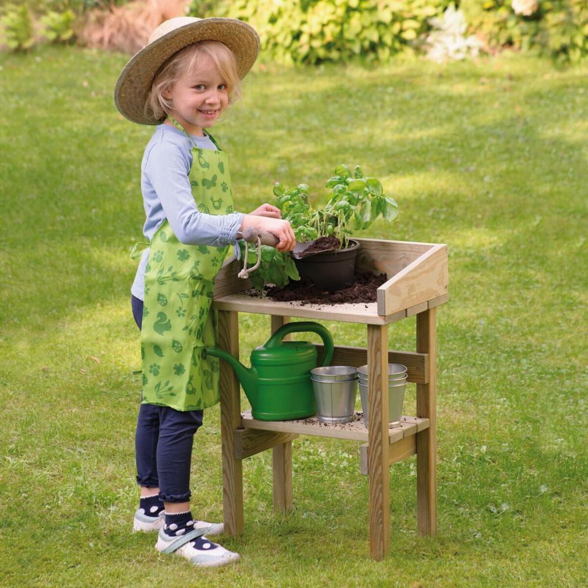 Ein Kind in Gartenkleidung steht vor einem Kinder-Pflanztisch und topft eine Pflanze ein. Der Pflanztisch besteht aus hellem Holz und hat zwei Etagen, auf denen sich weitere Pflanzenbehälter und Gartengeräte befinden. Im Hintergrund ist eine grüne Rasenfläche zu sehen.