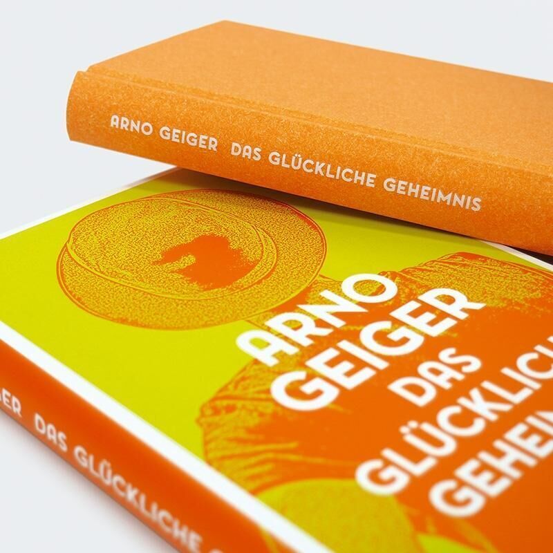 Das Buch 'Das glückliche Geheimnis' von Arno Geiger zeigt ein Cover mit dominierenden Farben Orange und Gelb. Der Titel und der Name des Autors sind in Weiß aufgedruckt.