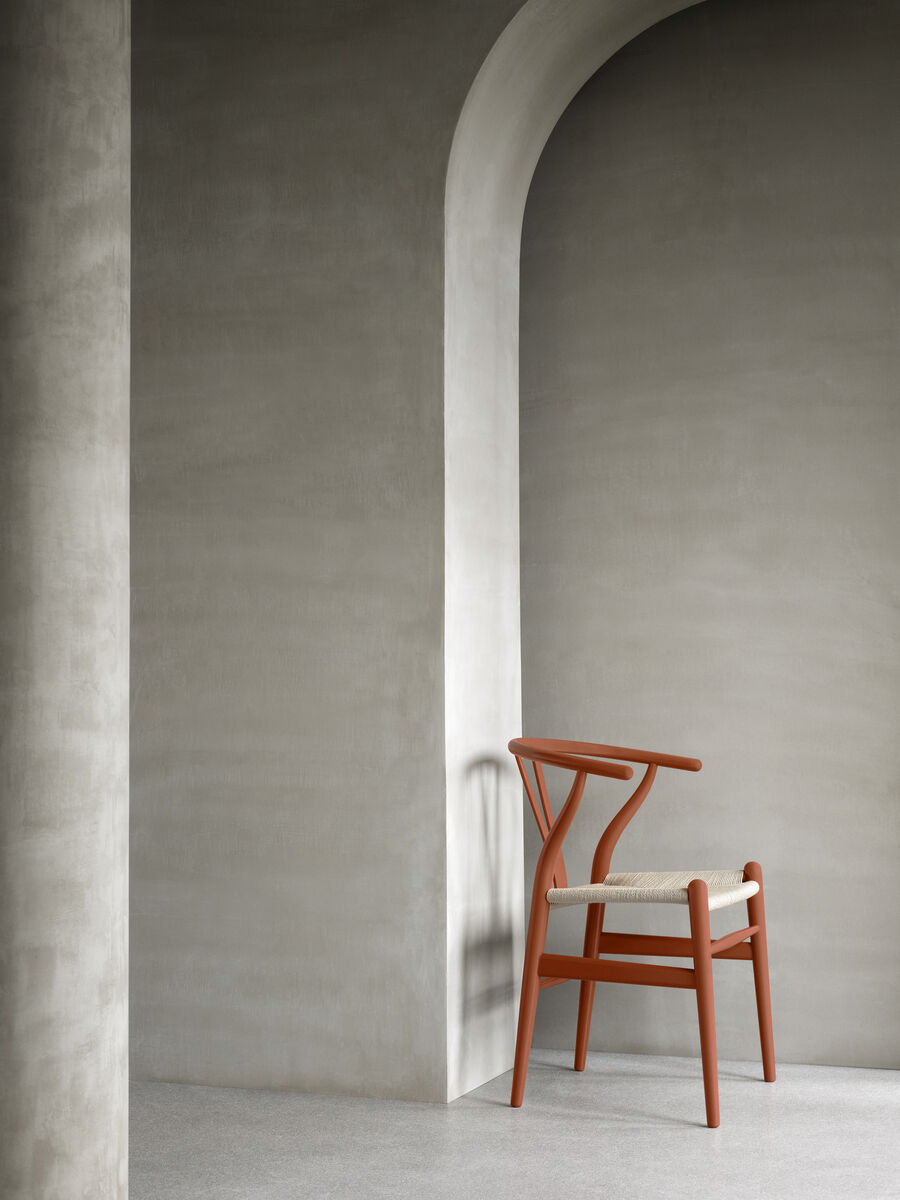 Ein Sonderedition »Stuhl CH24 Soft« von Carl Hansen. Der Stuhl hat eine minimalistische, skandinavische Designästhetik, ist aus braunem Holz gefertigt und verfügt über eine beige Sitzfläche. Der Hintergrund zeigt eine graue, futuristische Umgebung mit einem Bogen und einer Säule.