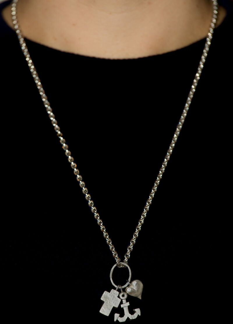 Nahaufnahme einer Kette mit Anhänger, die die Symbole Kreuz, Herz und Anker zeigt. Die Kette wird von einer Person getragen und der Hintergrund ist schwarz.