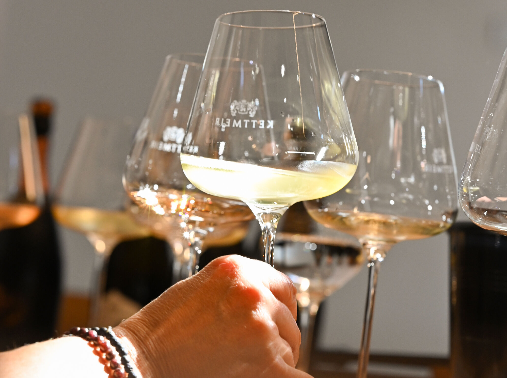 Hand hält ein Weinglas mit weißem Wein, weitere Weingläser stehen auf einem Tisch.