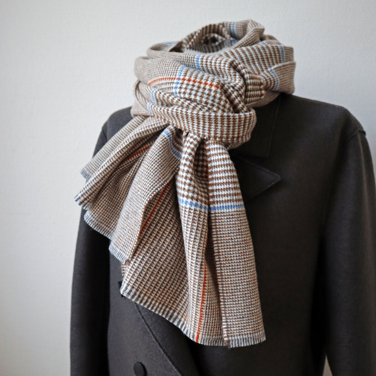 Merinoschal mit feinem Karomuster in Beige, Braun und blauen sowie orange Akzenten, stilvoll um den Kragen eines dunkelgrauen Mantels gelegt. Das Gesamtbild vermittelt einen eleganten, klassischen Look für die kalte Jahreszeit.