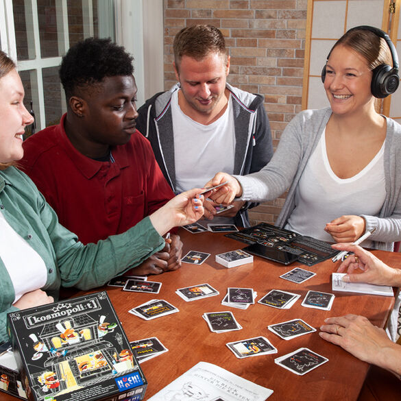 Fünf Personen sitzen gemeinsam am Tisch und spielen das Brettspiel Kosmopoli:t. Auf dem Tisch liegen Karten und Spielfiguren, während eine Person Kopfhörer trägt. Lächelnde Gesichter zeigen gemeinsame Freude am Spiel. Die Atmosphäre wirkt freundlich und interaktiv.