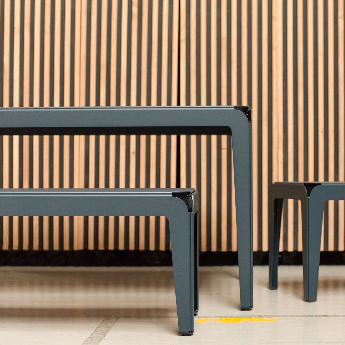Eine moderne Bank namens 'Bended Bench' aus Metall und Holz, dargestellt vor einer lamellierten Holzwand. Das Design ist minimalistisch und elegant.