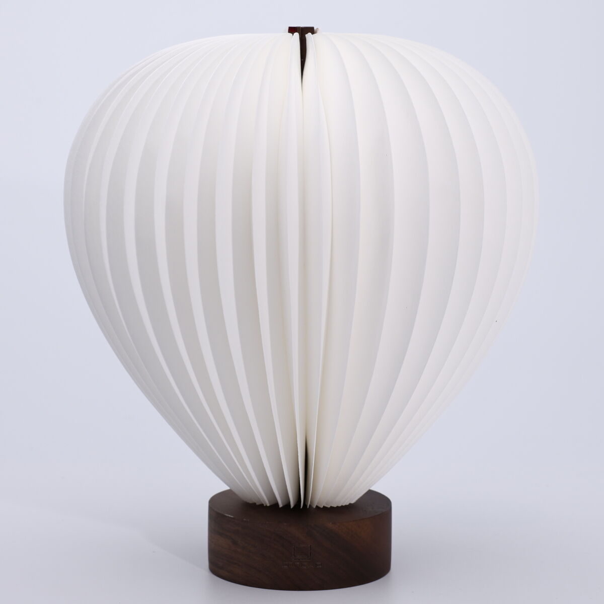 Die Leuchte »Aeris Light« von Gingko Design Ltd steht auf einer dunklen Holzbasis aus Walnussholz. Ihr Körper ist ballonförmig, besteht aus vielen gefächerten weißen Lamellen und erinnert an einen Heißluftballon. Der Hintergrund ist neutral hellgrau.