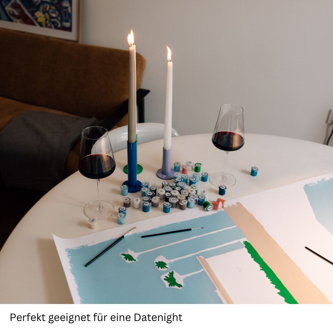Auf dem Bild sehen wir ein Malen-nach-Zahlen-Set mit dem Titel 'Schmeckt Oysters Gut', das auf einem weißen Tisch ausgebreitet ist. Zwei Weingläser mit rotem Wein und zwei angezündete weiße und blaue Kerzen stehen neben dem Set. Der Hintergrund des Bildes zeigt ein braunes Sofa und ein abstraktes Gemälde an der Wand. Die Szene wirkt sehr romantisch und lädt zur Entspannung ein.