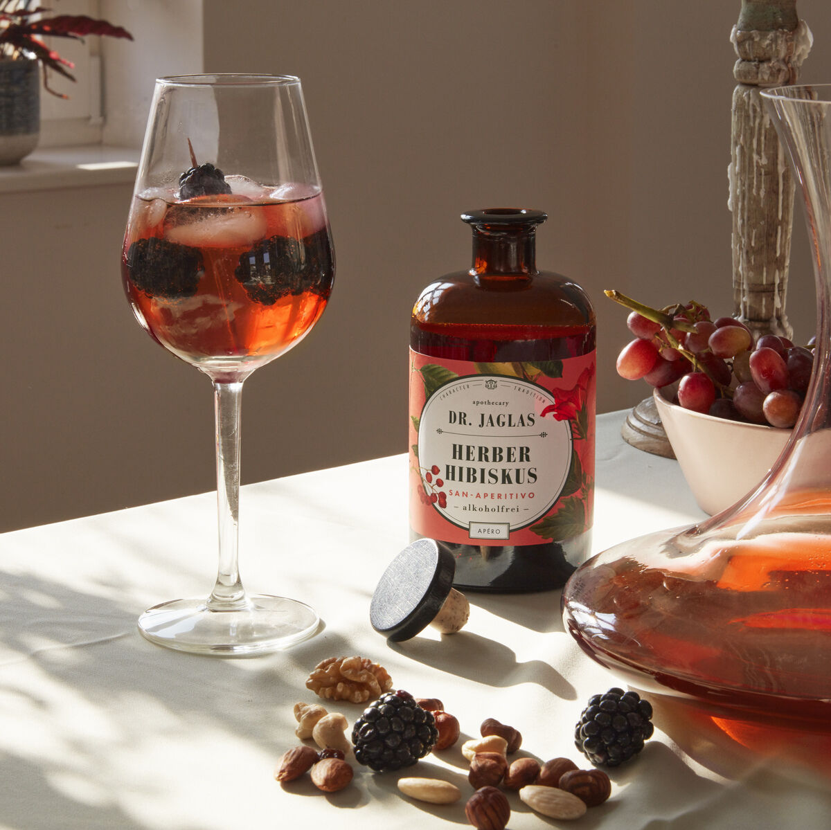 Ein Glas mit Herber Hibiskus Aperitif, verziert mit Eis und Früchten, steht auf einem Tisch neben einer Flasche der nicht-alkoholischen Getränks. Nüsse und Früchte sind ebenfalls auf dem Tisch verstreut. Im Hintergrund sind Dekor und Fensterlicht sichtbar.
