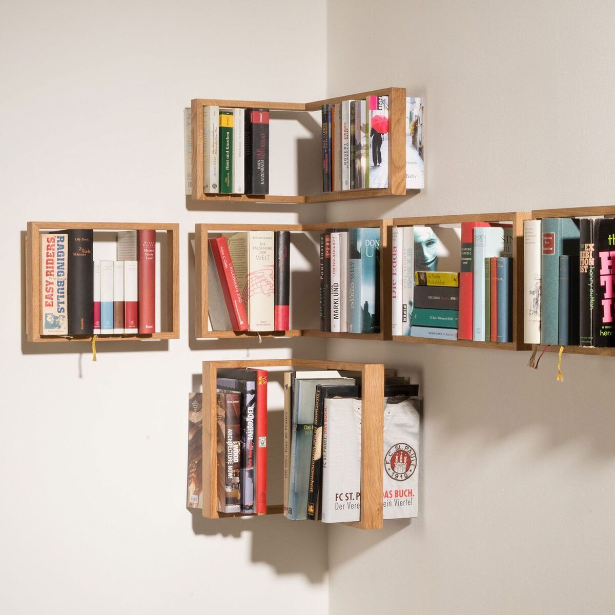 Ein modernes Holzregal namens „b-eck“ von Regal, das an einer weißen Wand angebracht ist. Es besteht aus sechs kleinen Regaleinheiten, in denen verschiedene Bücher in verschiedenen Farben und Größen aufbewahrt werden.