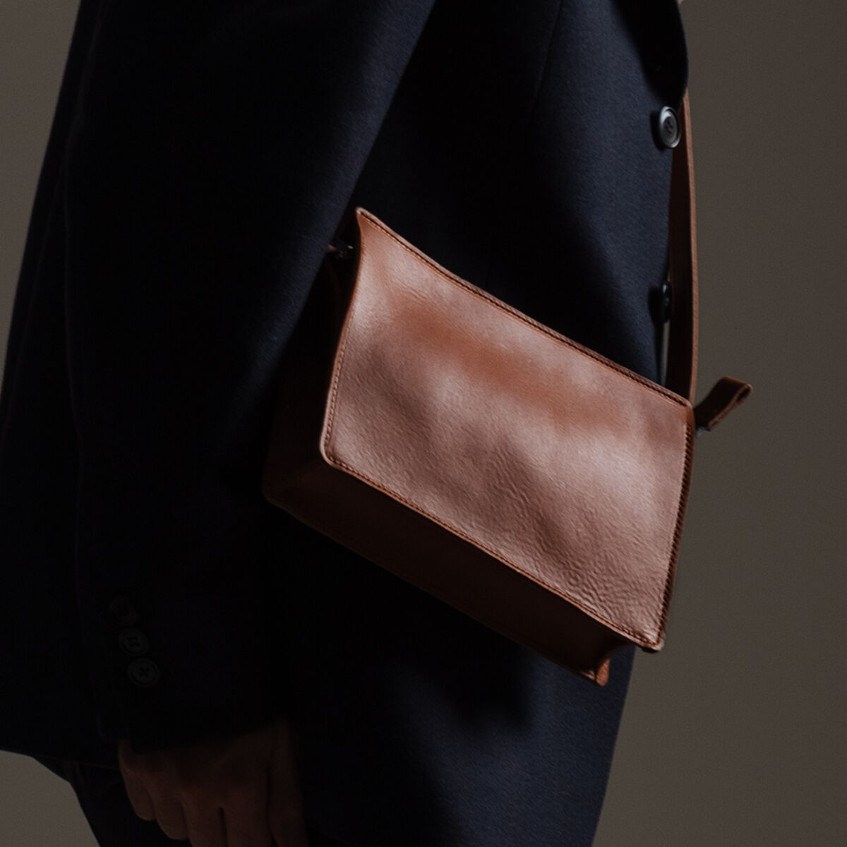 Eine rechteckige Umhängetasche aus glattem, braunem Kernleder wird von einer Person in dunkler Kleidung seitlich über der Schulter getragen. Die Tasche hat ein minimalistisches, elegantes Design und ist hochwertig verarbeitet. Das Bild ist dezent und im Studio mit neutralem Hintergrund aufgenommen.