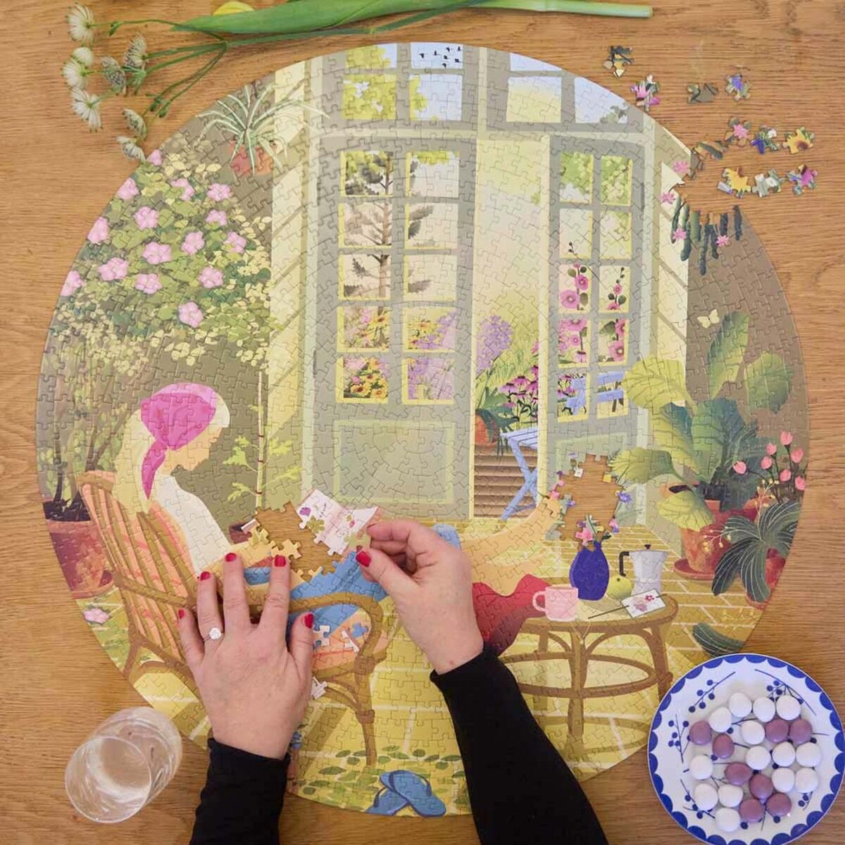 Rundes 1000-Teile Puzzle »Morning Glow« von VISSEVASSE zeigt eine idyllische Morgenszene mit einer sitzenden Frau im Wintergarten, umgeben von Zimmerpflanzen, Fenstern und weichem Sonnenlicht. Eine Person setzt das Puzzle zusammen. Auf dem Holztisch liegen Blumen und eine Schale mit Süßigkeiten.