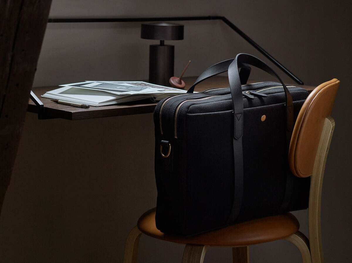 Schwarze Businesstasche ENDEAVOUR von Mismo auf einem Stuhl, neben einem modernen Schreibtisch mit Büchern und einer Lampe.
