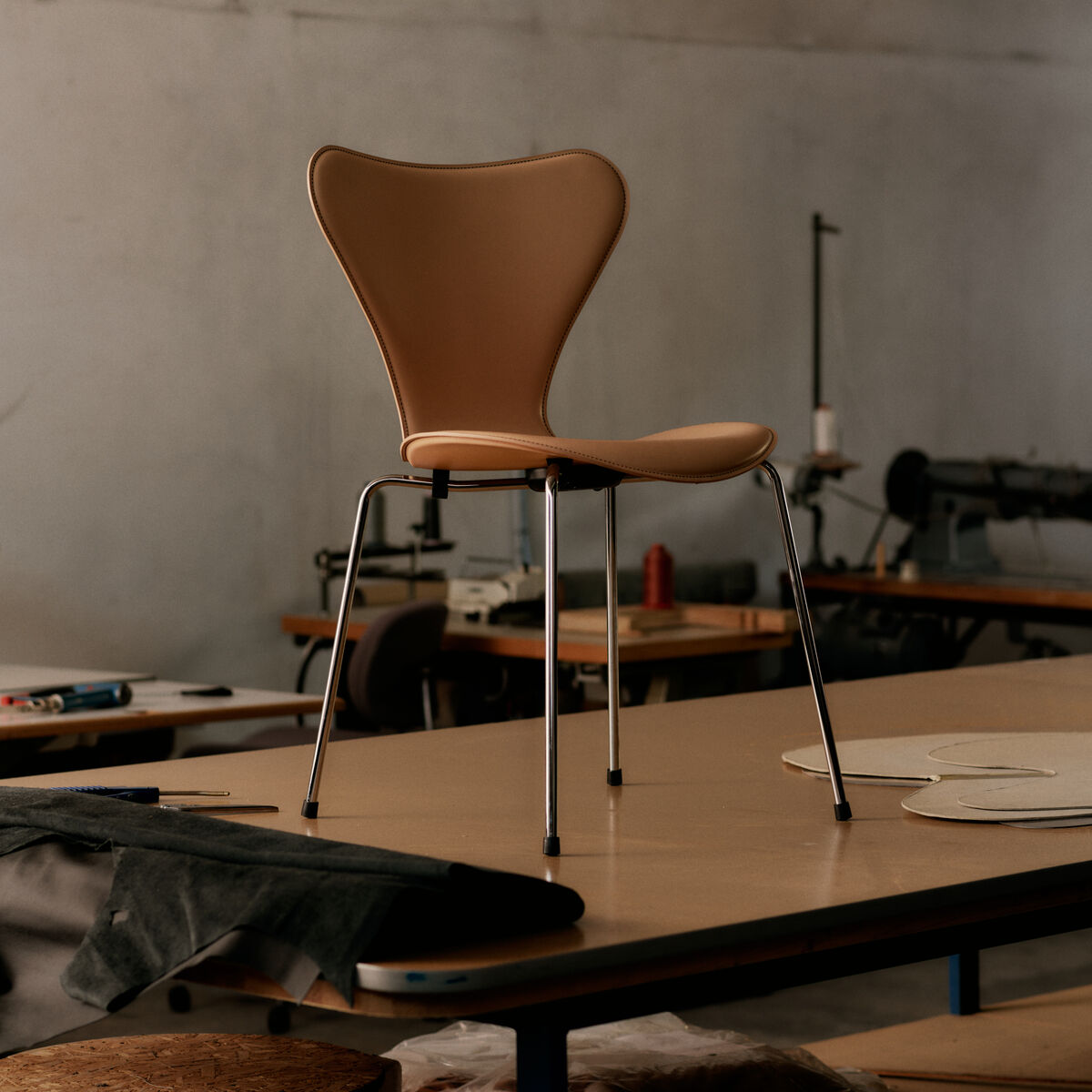 Der Stuhl »Series 7™ Tailored« von Fritz Hansen steht zentral auf einem Tisch in einer Werkstattumgebung. Der Stuhl zeigt eine elegante, geschwungene Sitzfläche in warmem Ecru-Ton und glänzende Chrombeine. Im Hintergrund sind Tische, Stoffe und Industrie-Nähmaschinen sichtbar.