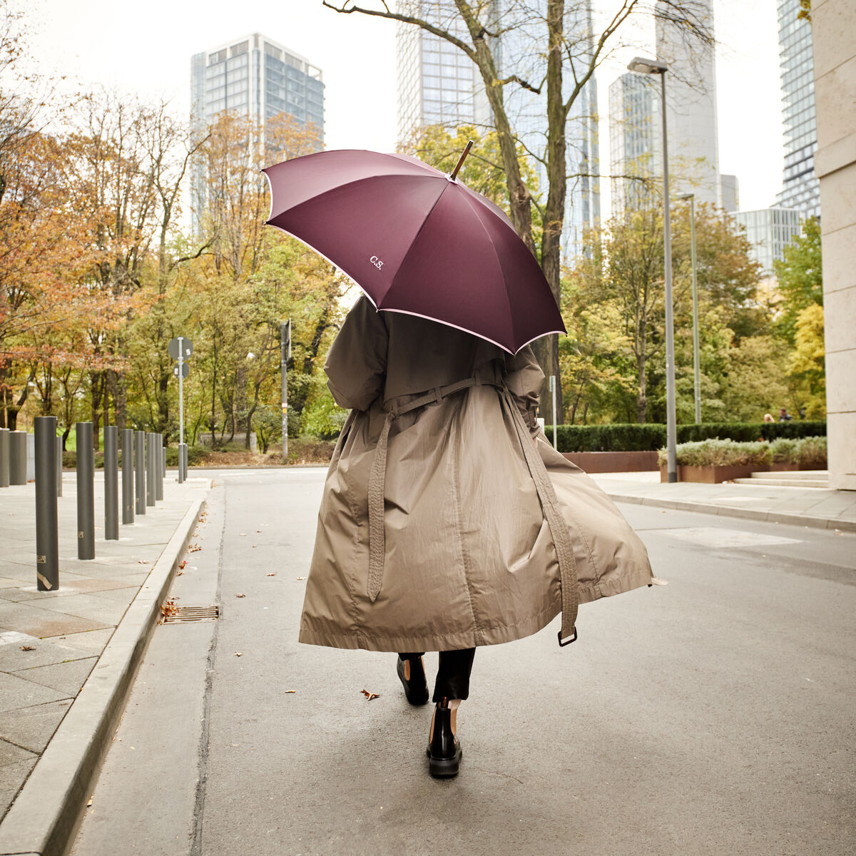Eine Frau spaziert in einer beigen Trenchcoat bei herbstlichem Wetter durch eine städtische Umgebung und hält einen Beeren-farbenen Regenschirm 'Norfolk Oxford'. Hochhäuser und herbstliche Bäume sind im Hintergrund zu sehen.