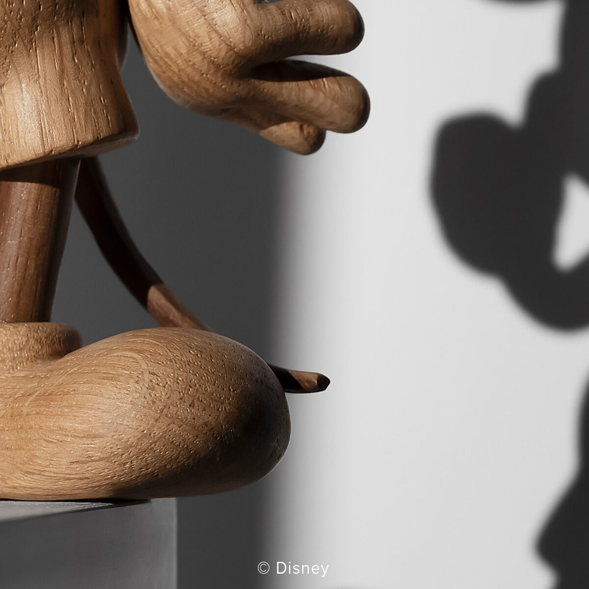 Detailreiche Holzfigur „Mickey Maus“ von Boyhood, gefertigt aus europäischer Eiche, mit ausgestrecktem Arm, auf weißem Podest. Im Hintergrund deutlich sichtbarer, charakteristischer Schatten der Figur auf grauer Wand. Modernes, minimalistisches Design.