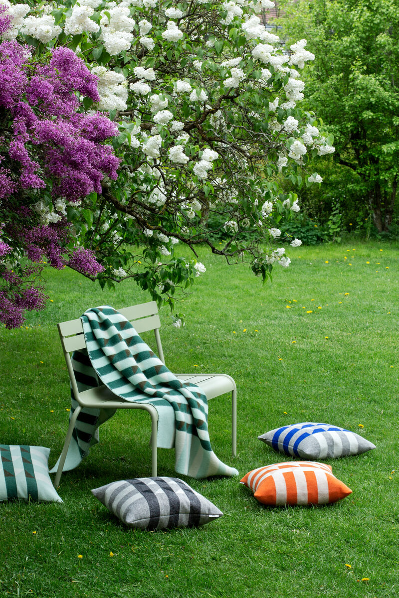 Eine Gartenlandschaft mit mehreren farbenfrohen Streifenkissen und einer Decke auf einem grünen Stuhl. Im Hintergrund blühende Büsche und eine grüne Wiese.