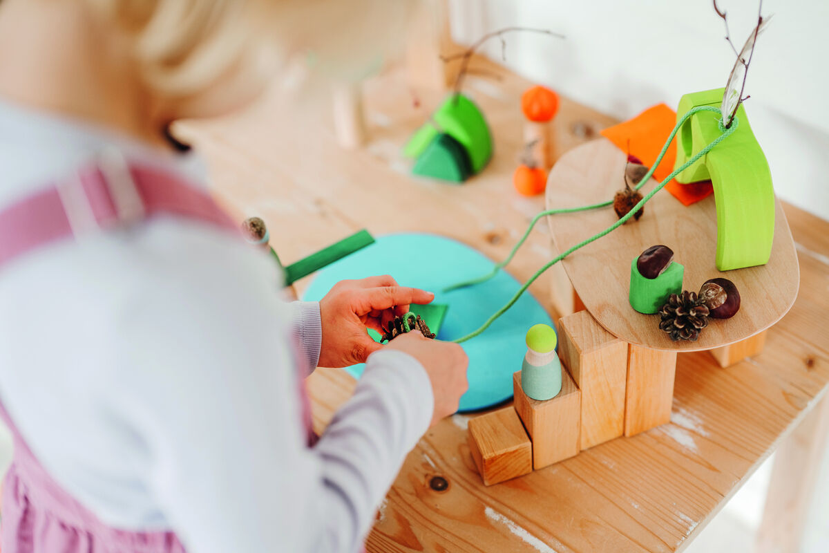 Ein Kind spielt mit einem Spielzeugset namens 'Spielwelt Waldlichtung'. Die bunten Holzbausteine in Grün, Orange und Blau darstellen eine Waldszene auf einem Holztisch. Das Set umfasst auch natürliche Elemente wie Kiefernzapfen und Zweige.