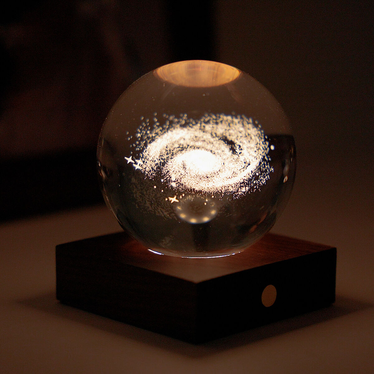 Eine dekorative Lampe namens »Amber Light«, bestehend aus einer klaren Glaskugel mit einem leuchtenden Galaxie-Muster im Inneren. Die Kugel ruht auf einem quadratischen Holzsockel und erzeugt eine warme, stimmungsvolle Beleuchtung, die sich ideal als Wohnaccessoire oder Geschenk eignet.