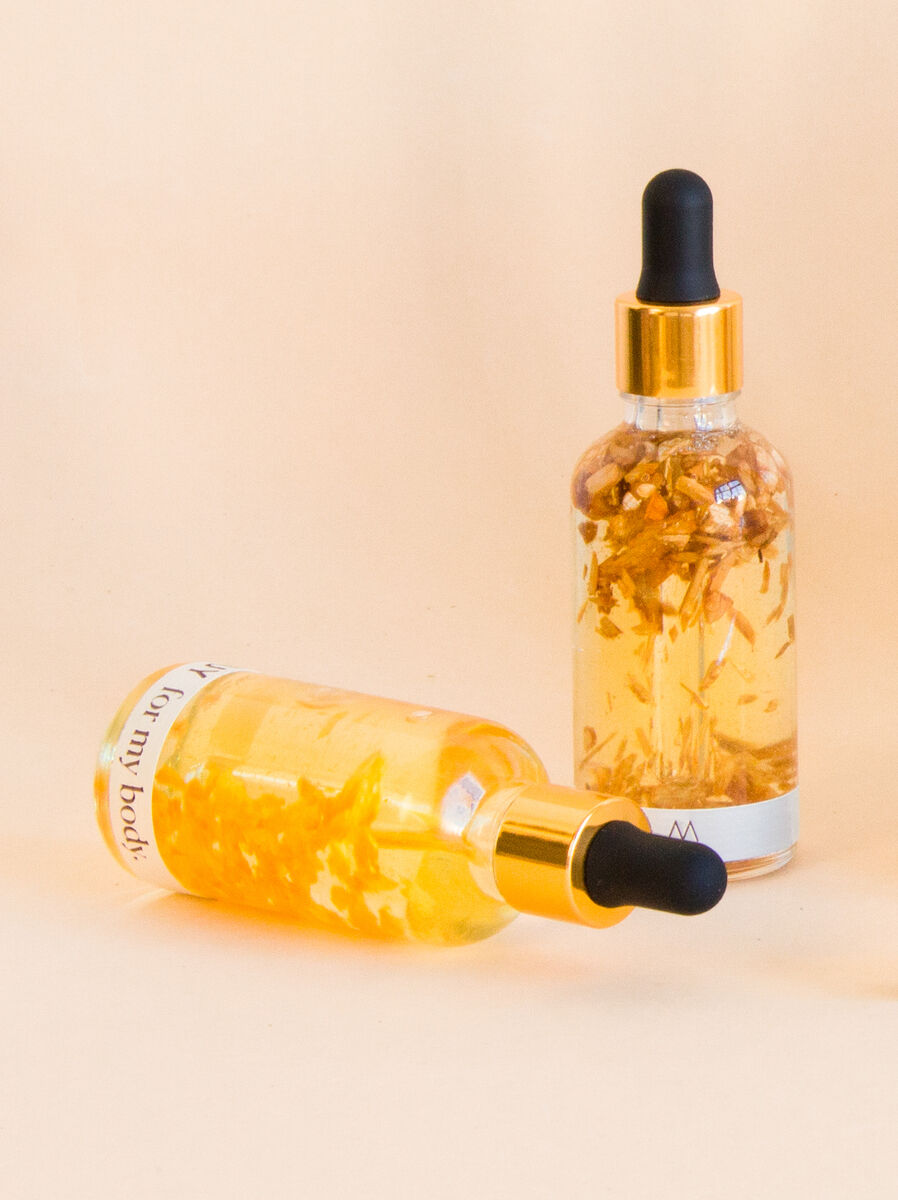 Das Bild zeigt ein Naturkosmetik-Set der Marke MUY. Es besteht aus zwei Glasflaschen mit goldfarbenen Verschlüssen und schwarzem Pipettenaufsatz. Die Flaschen enthalten eine gelbe Flüssigkeit und Blütenextrakte. Die Hintergrundfarbe ist cremefarben.