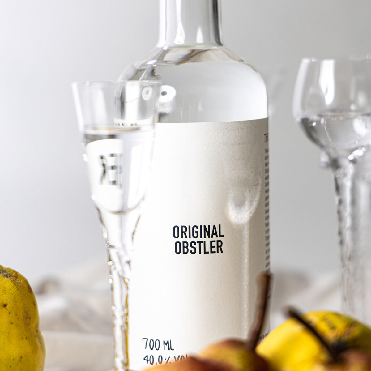 Eine Flasche Obstler »HAINBER« von AASPIRITS, 700 ml, mit weißem, minimalistischen Etikett steht neben zwei Gläsern und frischem Obst wie Apfel und Birne. Die Szene ist hell ausgeleuchtet, der Hintergrund unscharf und lichtdurchflutet.