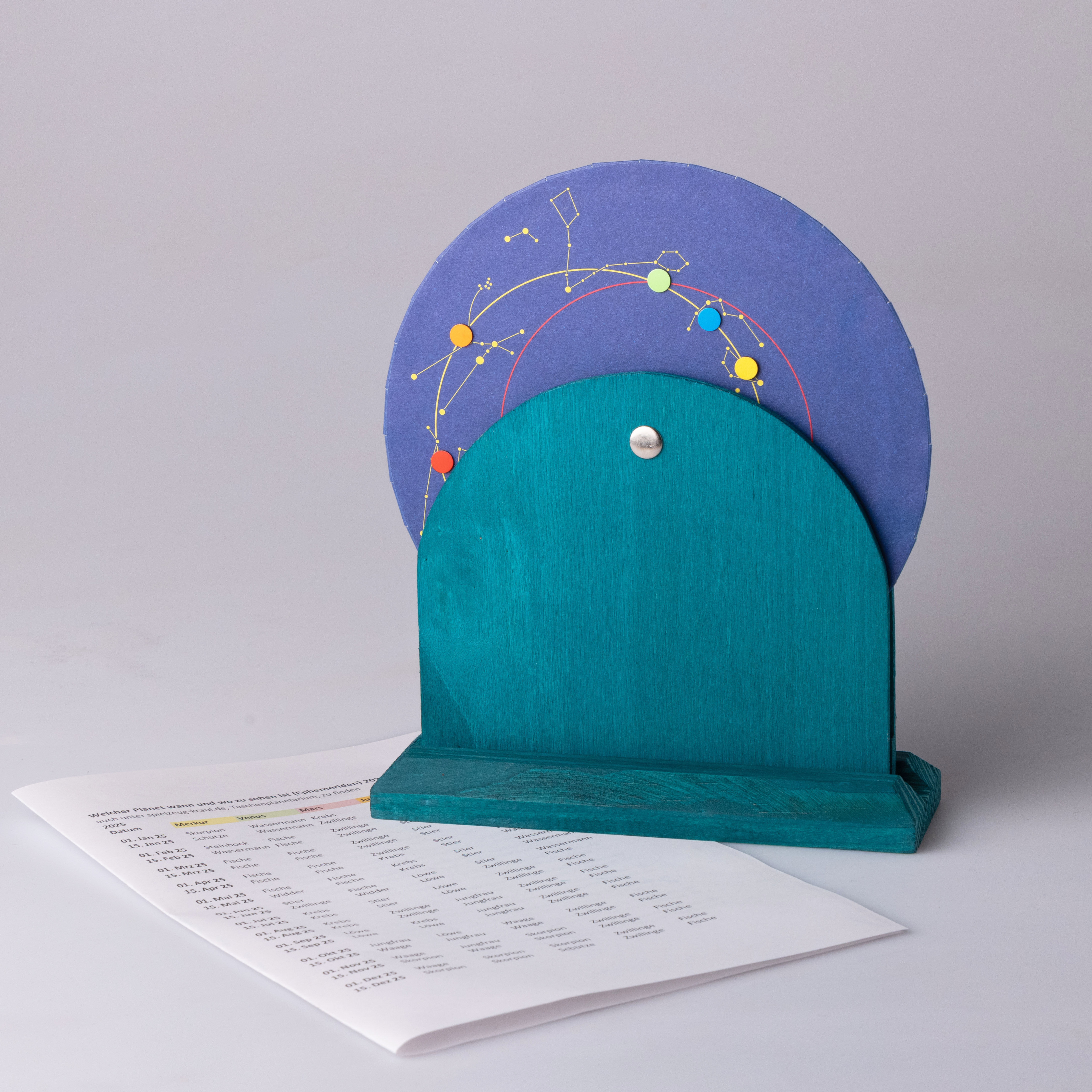 Ein Taschenplanetarium mit einem türkisfarbenen Holzsockel und einer drehbaren, lila Scheibe mit farbigen Markierungen, auf der Sternbilder und Himmelsbahnen dargestellt sind. Im Hintergrund liegt ein Blatt Papier mit einer Liste bunter Textzeilen auf einem hellen Untergrund.