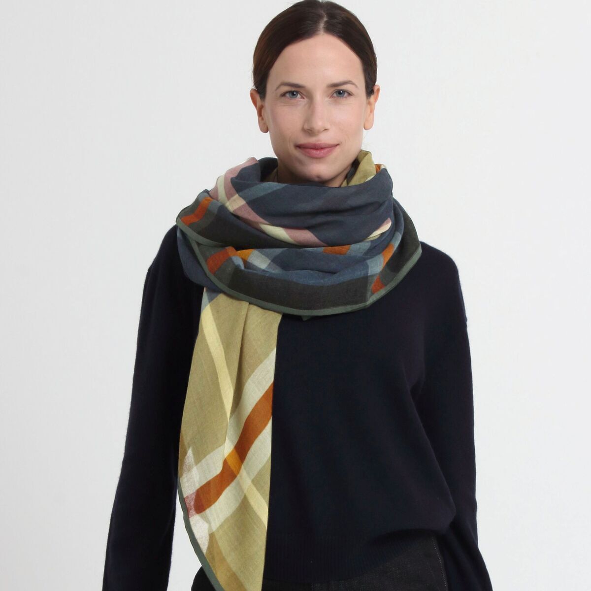 Frau trägt den Wollschal »Louise« Grün von Shalls Barcelona, 100 % Schurwolle, mit großflächigem Karomuster in Grün-, Beige-, Orange- und Grautönen. Schal locker um den Hals gewickelt, Outfit dunkel und schlicht, Hintergrund hell und neutral.