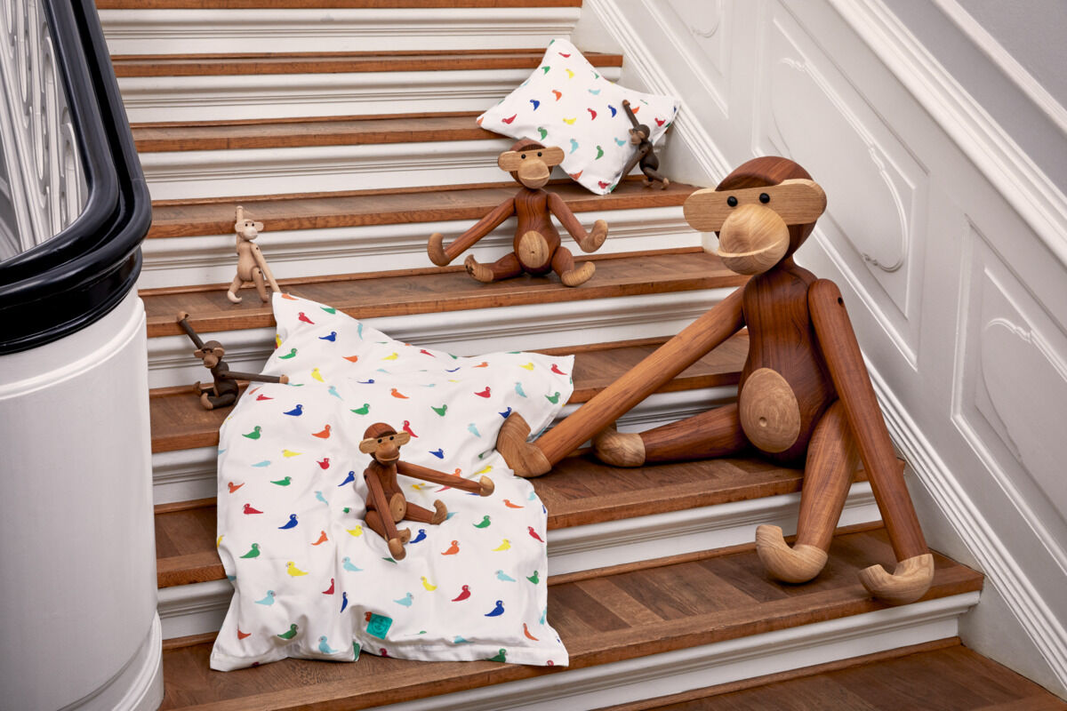 Mehrere Holzfiguren in Form von Affen sitzen auf einer Treppe. Die Figuren sind aus braunem Holz und befinden sich neben weißen Kissen mit bunten Mustern.