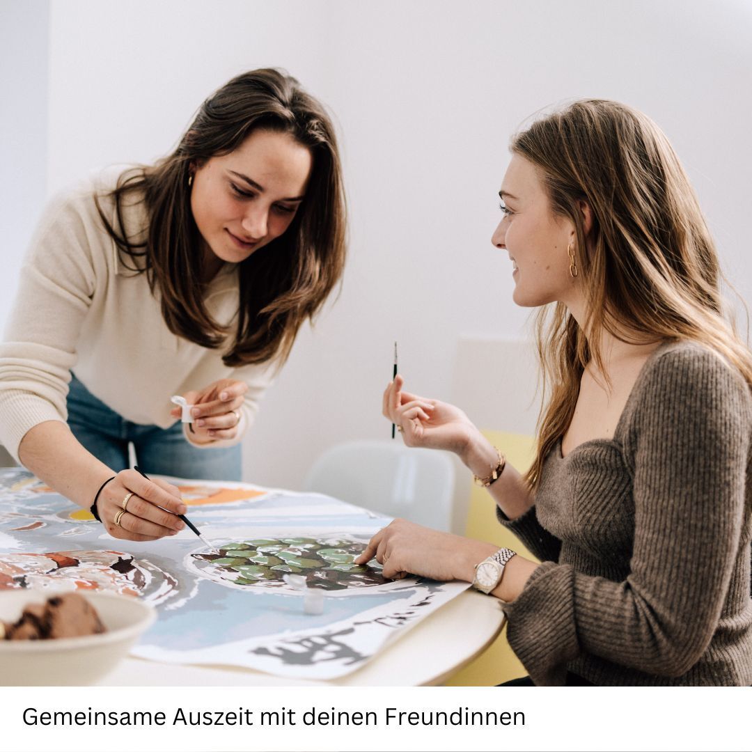 Zwei junge Frauen sitzen an einem Tisch und arbeiten zusammen an einem Malen nach Zahlen Projekt. Beide konzentrieren sich auf das Kunstwerk vor ihnen und scheinen Spaß an der Tätigkeit zu haben. Der Hintergrund ist in hellen Farben gehalten und auf dem Tisch liegen Malutensilien und Snacks.