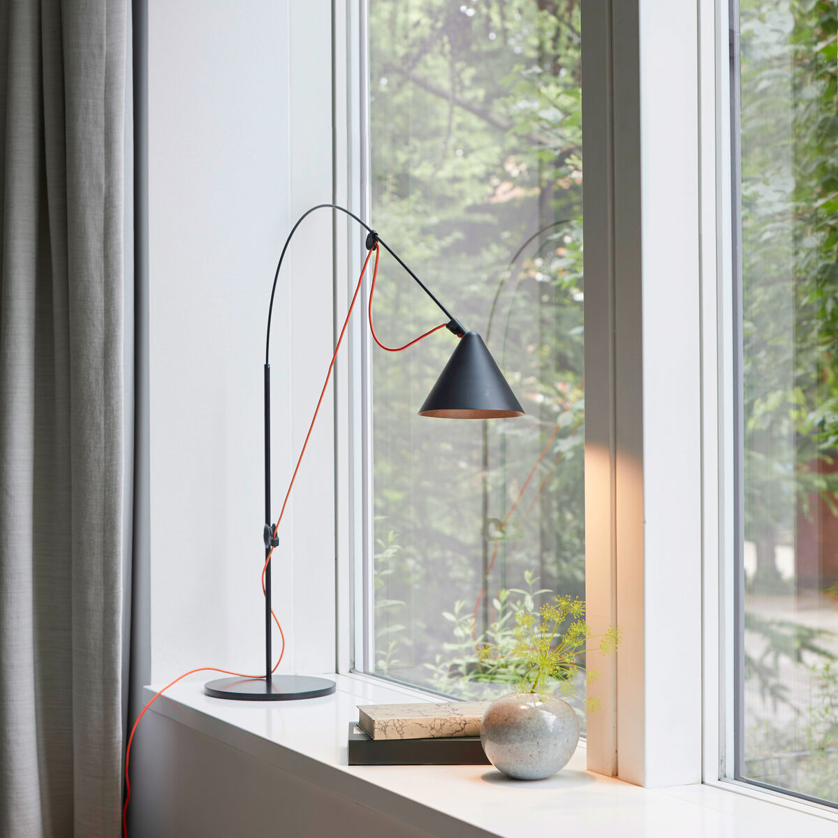 Eine moderne Tischlampe mit schwarzem Sockel und schwarzem, verstellbarem Arm, montiert auf einer Fensterbank. Der obere Lampenarm ist rot umwickelt. Die Lampe steht neben einem kleinen Topf mit Pflanzen und einem Buch. Im Hintergrund sind ein Fenster und ein grauer Vorhang zu sehen.