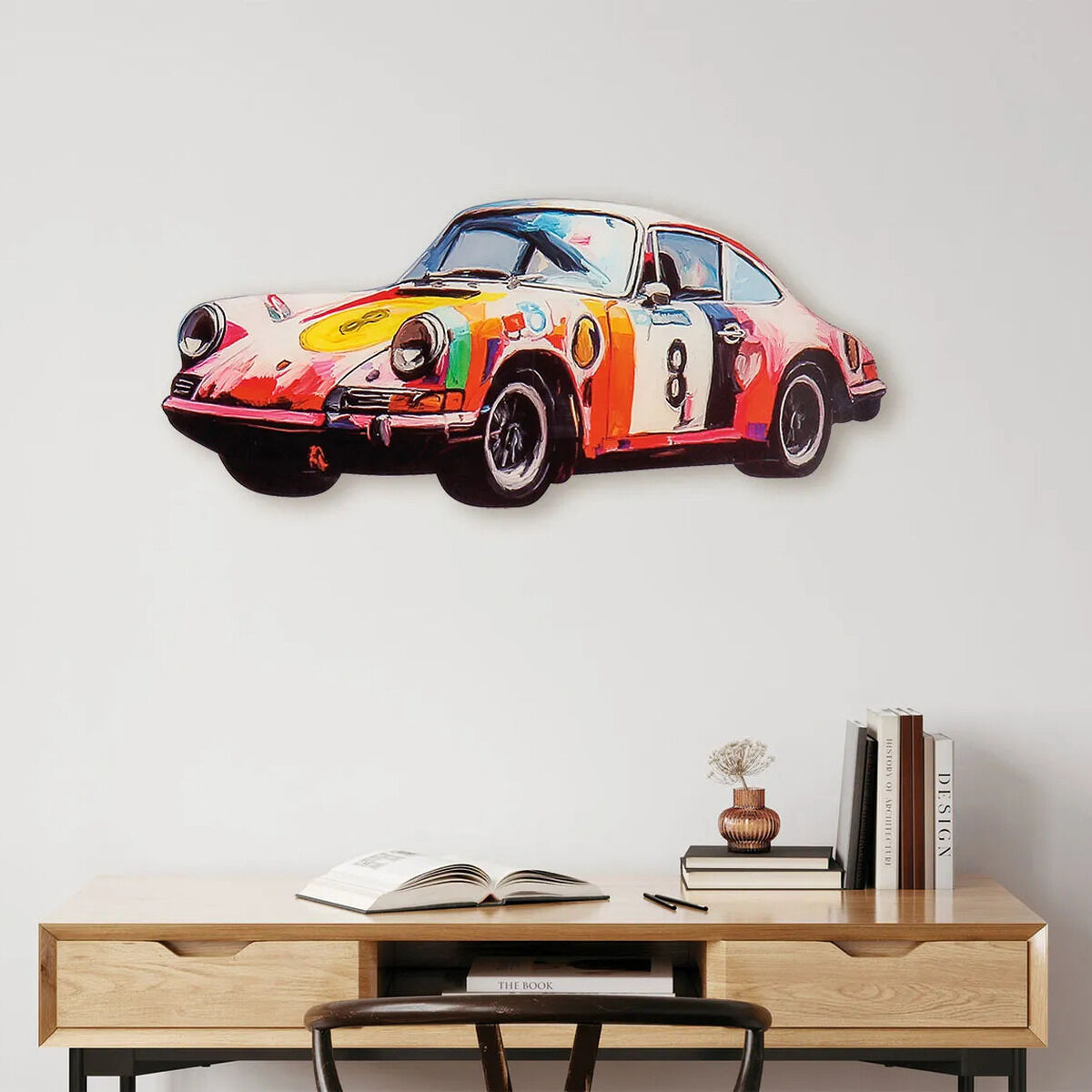 Auf dem Bild ist ein dekorativer Wandschmuck in Form eines sportlichen Porsche 911, gestaltet als farbenfrohes und detailreiches Cut-Out, zu sehen. Das Auto weist bunte Akzente in Rot, Gelb, Orange und weiteren Farben auf. Der Porsche hängt über einem hellen Schreibtisch mit geöffnetem Buch und stilvollen Deko-Objekten.