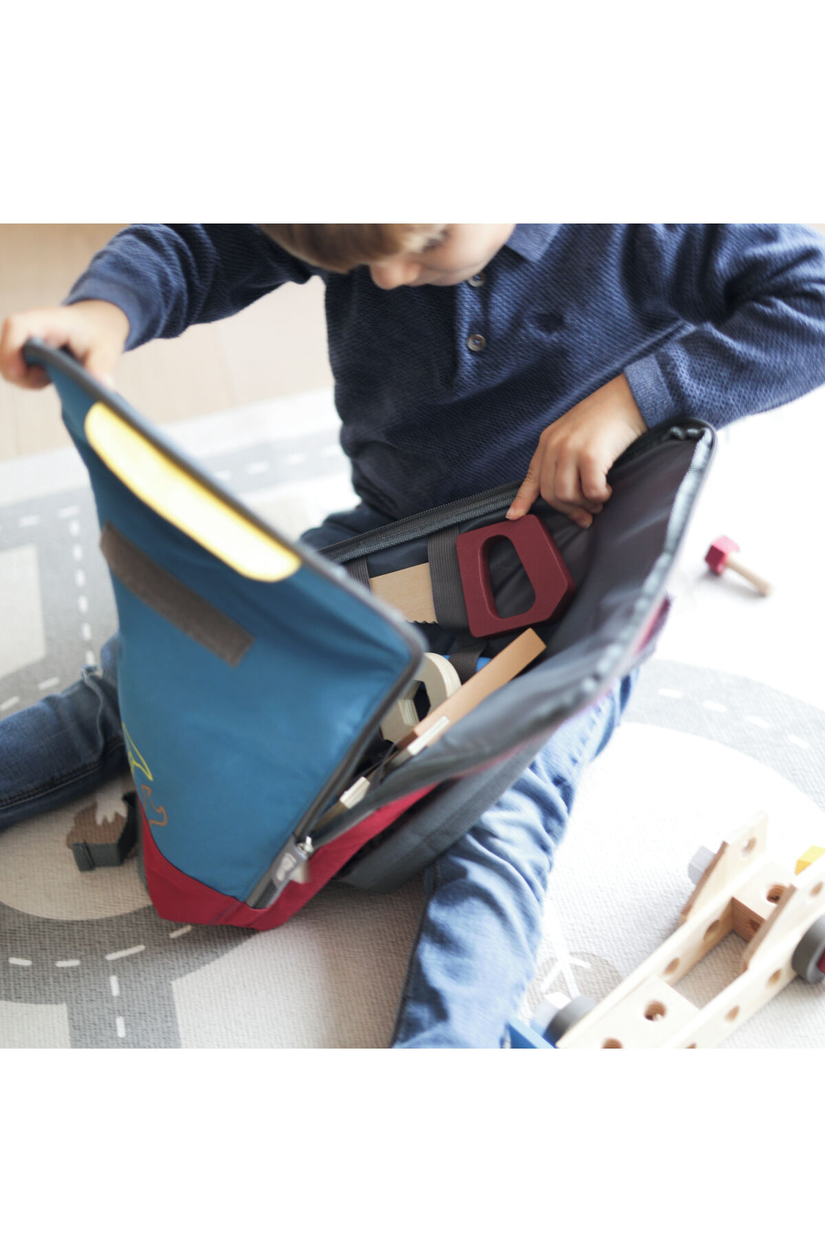 Ein Kind sitzt auf einem Teppich und spielt mit einem offenen Werkzeugrucksack, der verschiedene Holzspielzeuge und Werkzeugteile enthält. Der Rucksack ist blau und hat bunte Akzente. Neben dem Kind liegen weitere Bauteile.