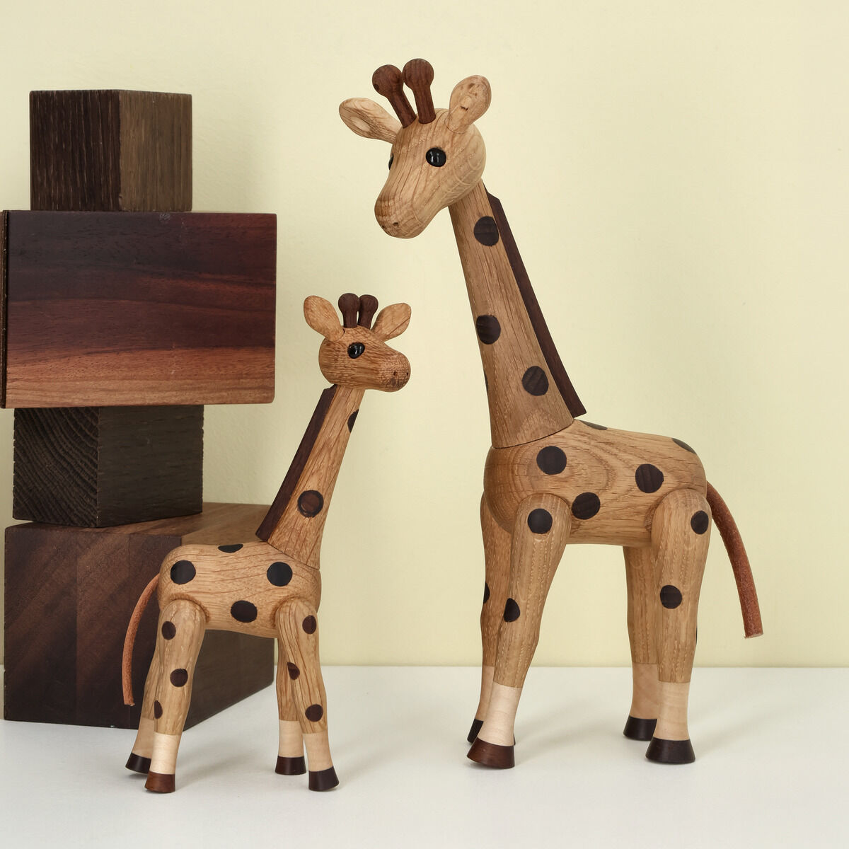 Zwei Holzfiguren einer Giraffe, Größe 'Klein' (Produktname: Giraffe, Artikelnummer: 52324), stehen nebeneinander auf einer weißen Oberfläche. Im Hintergrund stehen aus Holzklötzen gestapelte Würfel vor einer hellgelben Wand. Die Giraffen haben dunkle Punkte und detaillierte Details.