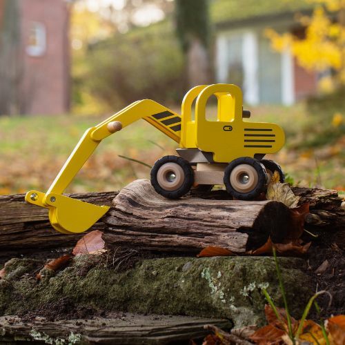 Gelber Holzbagger als Spielzeug steht auf einem Baumstamm im Freien. Das detailreiche Baufahrzeug ist aus Holz gefertigt und für Kinder geeignet. Im Hintergrund sind herbstliche Farben, Gras und unscharfe Gebäude sichtbar.