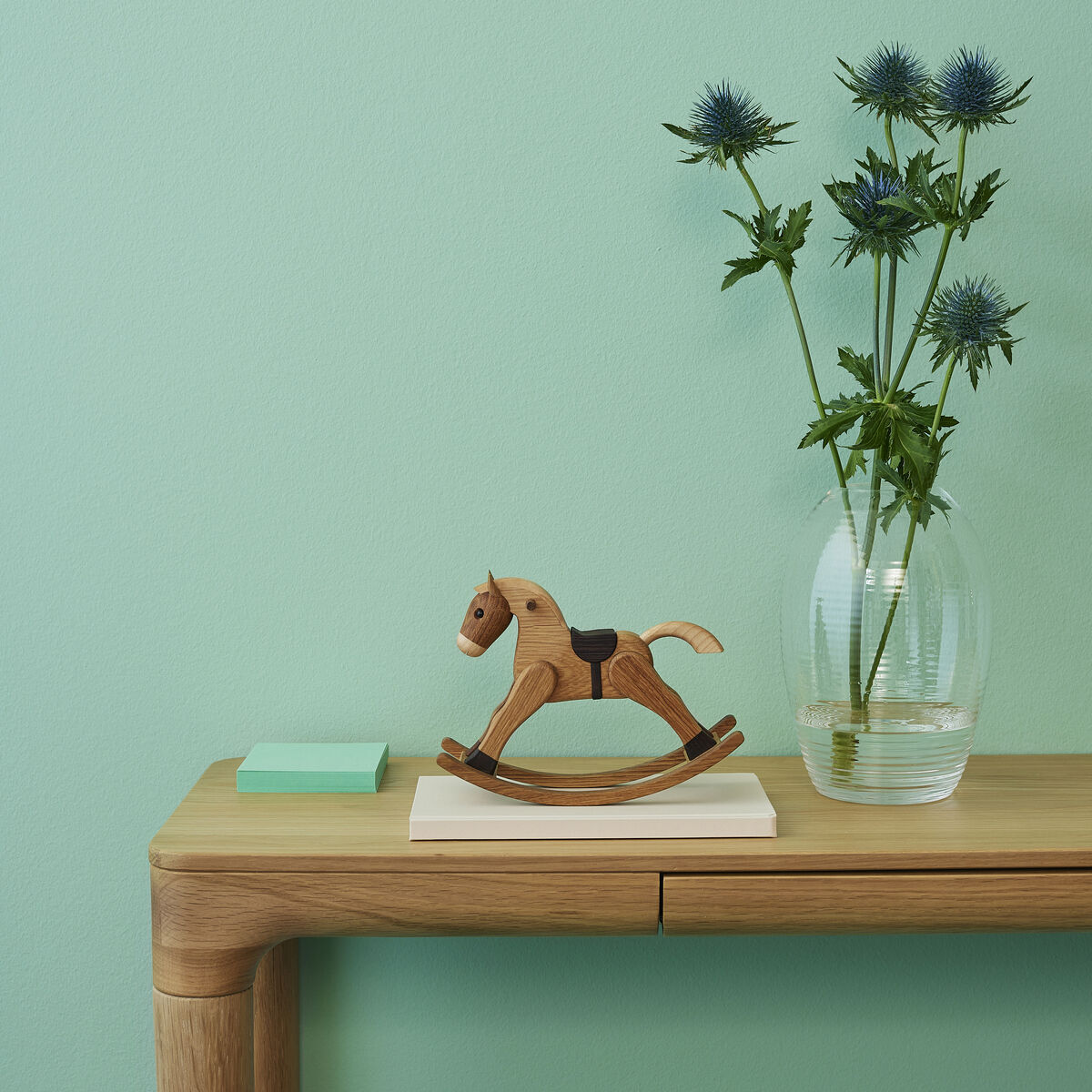 Eine dekorative Holzfigur in Form eines Schaukelpferds steht auf einem hellen Holztisch vor einer mintgrünen Wand. Daneben befindet sich eine Glasvase mit Distelblumen sowie ein kleiner, hellgrüner Block. Die Szene wirkt ruhig und minimalistisch.