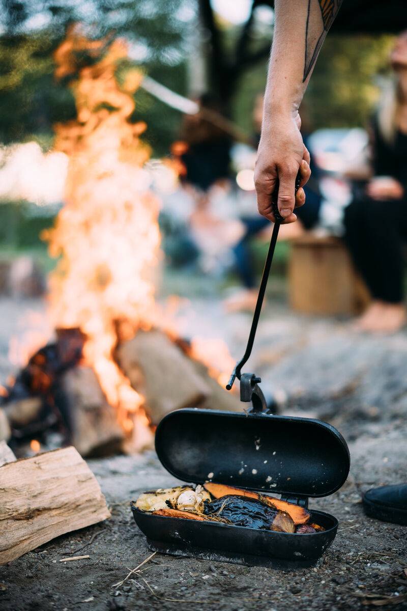 Eine Hand hält einen Kartoffelbräter aus Gusseisen über ein offenes Lagerfeuer. Im Hintergrund sind unscharf Menschen zu sehen, die die warme Atmosphäre genießen. Der Kartoffelbräter enthält mehrere gebratene Kartoffeln.
