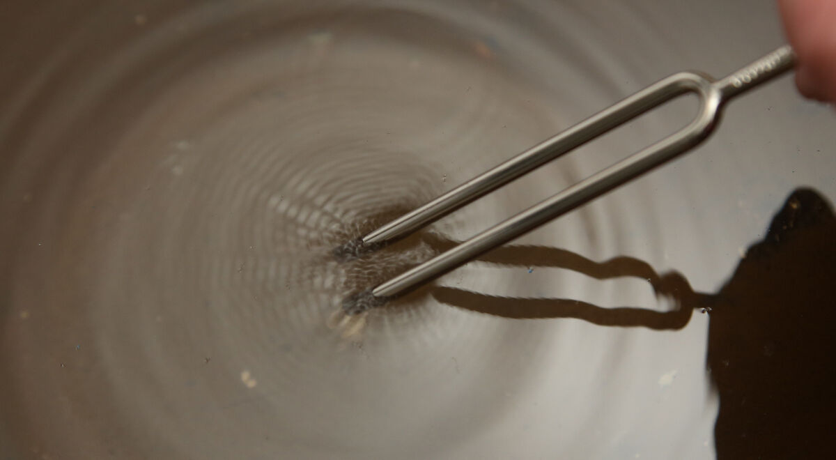 Nahaufnahme einer Stimmgabel, die in Wasser getaucht ist und Wellen erzeugt. Die Reflektion der Gabel ist auf der Wasseroberfläche sichtbar.