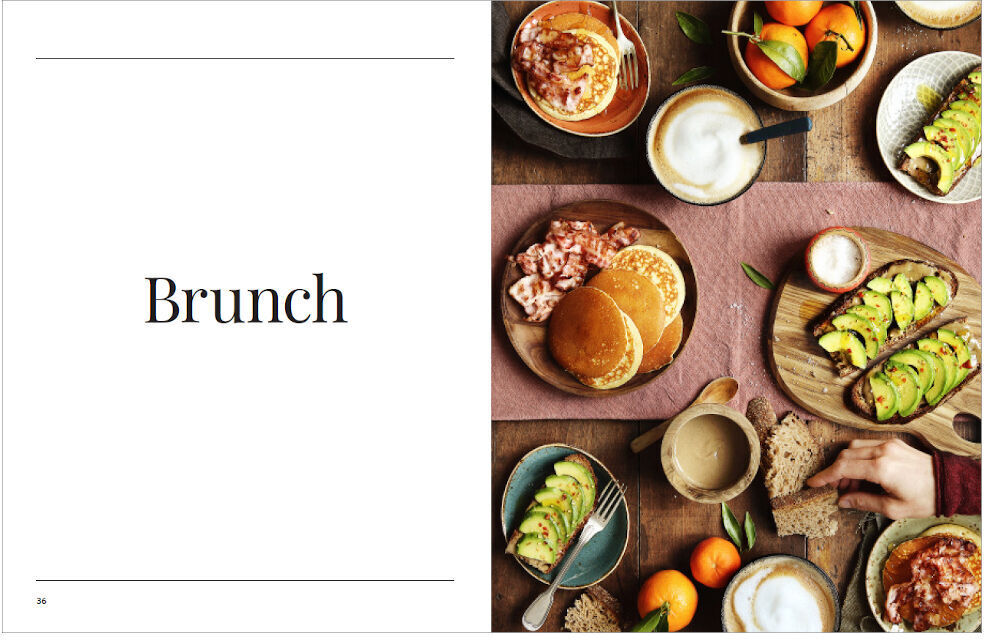 Ein aufgeschlagenes Kochbuch mit dem Titel 'Sonntagsessen' zeigt auf der linken Seite das Wort 'Brunch'. Die rechte Seite zeigt eine Auswahl an Gerichten, darunter Pancakes mit Erdbeeren, Avocado-Toast, frisches Obst und Getränke. Der Tisch und die Teller haben warme Erdtöne.