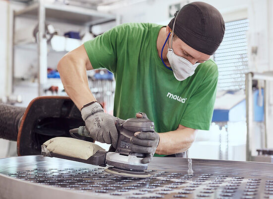 Ein Arbeiter in einem grünen Hemd bearbeitet in einer Werkstatt, umgeben von Werkzeugen und Maschinen, ein großes Blech mit einer elektrischen Schleifmaschine.