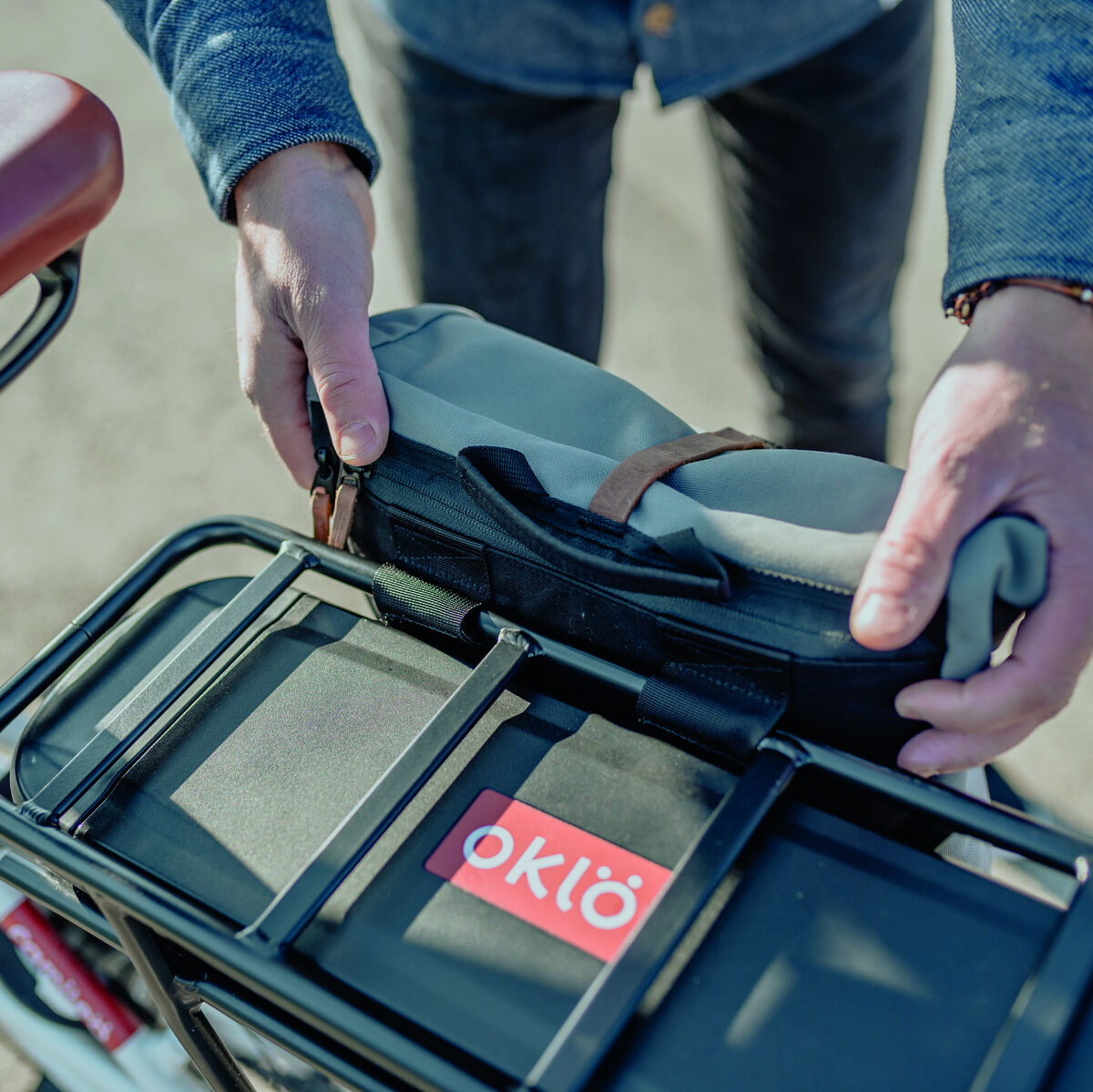 Fahrradtasche »Annecy« wird an einem schwarzen Fahrradgepäckträger befestigt. Die Tasche ist teilweise grau und wird von zwei Händen gehalten. Es ist ein Fahrradrahmen und ein brauner Fahrradsattel sichtbar.