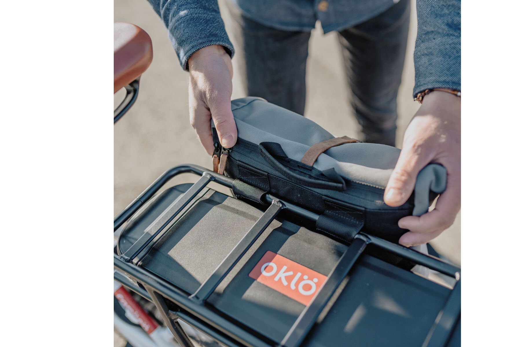 Fahrradtasche »Annecy« wird an einem schwarzen Fahrradgepäckträger befestigt. Die Tasche ist teilweise grau und wird von zwei Händen gehalten. Es ist ein Fahrradrahmen und ein brauner Fahrradsattel sichtbar.