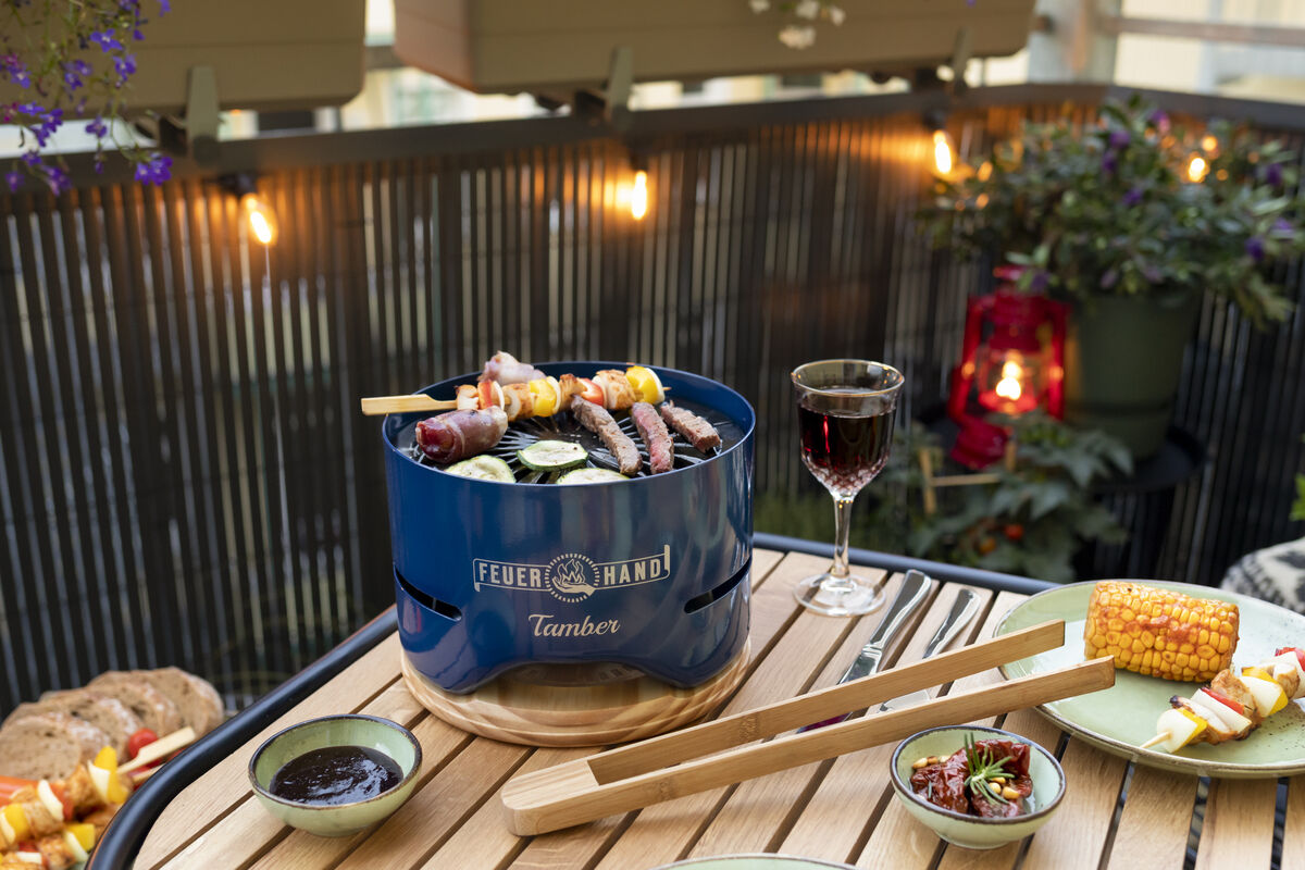 Tischgrill 'Tamber' von Feuerhand auf einem Holztisch im Freien, dekorativ dekoriert. Surrounding Grillbesteck, ein Glas Rotwein und gegrilltes Gemüse auf Tellern. Balkondekorationen und beleuchtete Lichter im Hintergrund.