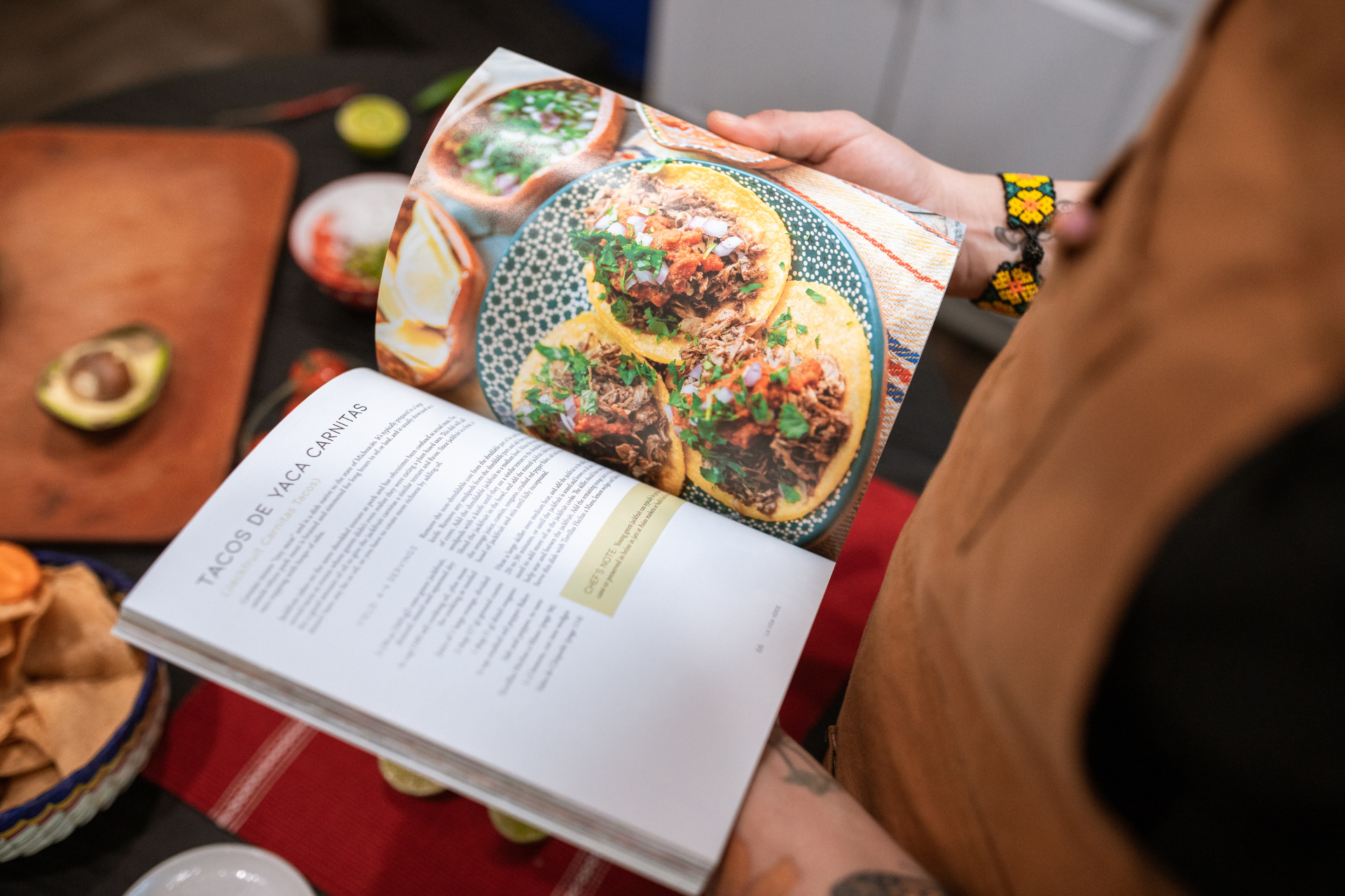 Link zur Kategorie Kochbücher. Eine Person hält ein aufgeschlagenes Kochbuch. Auf einer Seite sind drei fotografierte Tacos mit Fleischfüllung, Kräutern und gelben Tortillas auf gemustertem Teller abgebildet. Die andere Seite zeigt das Rezept für Tacos. Im Hintergrund liegen Zutaten wie Avocado und Chips auf Holzbrettern und einem roten Tuch.