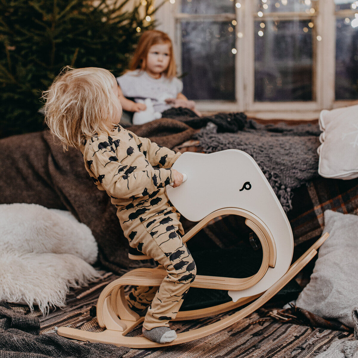 Ein kleines Kind schaukelt auf einem Schaukeltier in Elefantenform aus Holz in einem gemütlich dekorierten Wohnzimmer mit Decken, Kissen und Lichterketten. Im Hintergrund sitzt ein weiteres Kind vor einem festlichen Weihnachtsbaum. Das Bild vermittelt eine warme und familienfreundliche Atmosphäre.