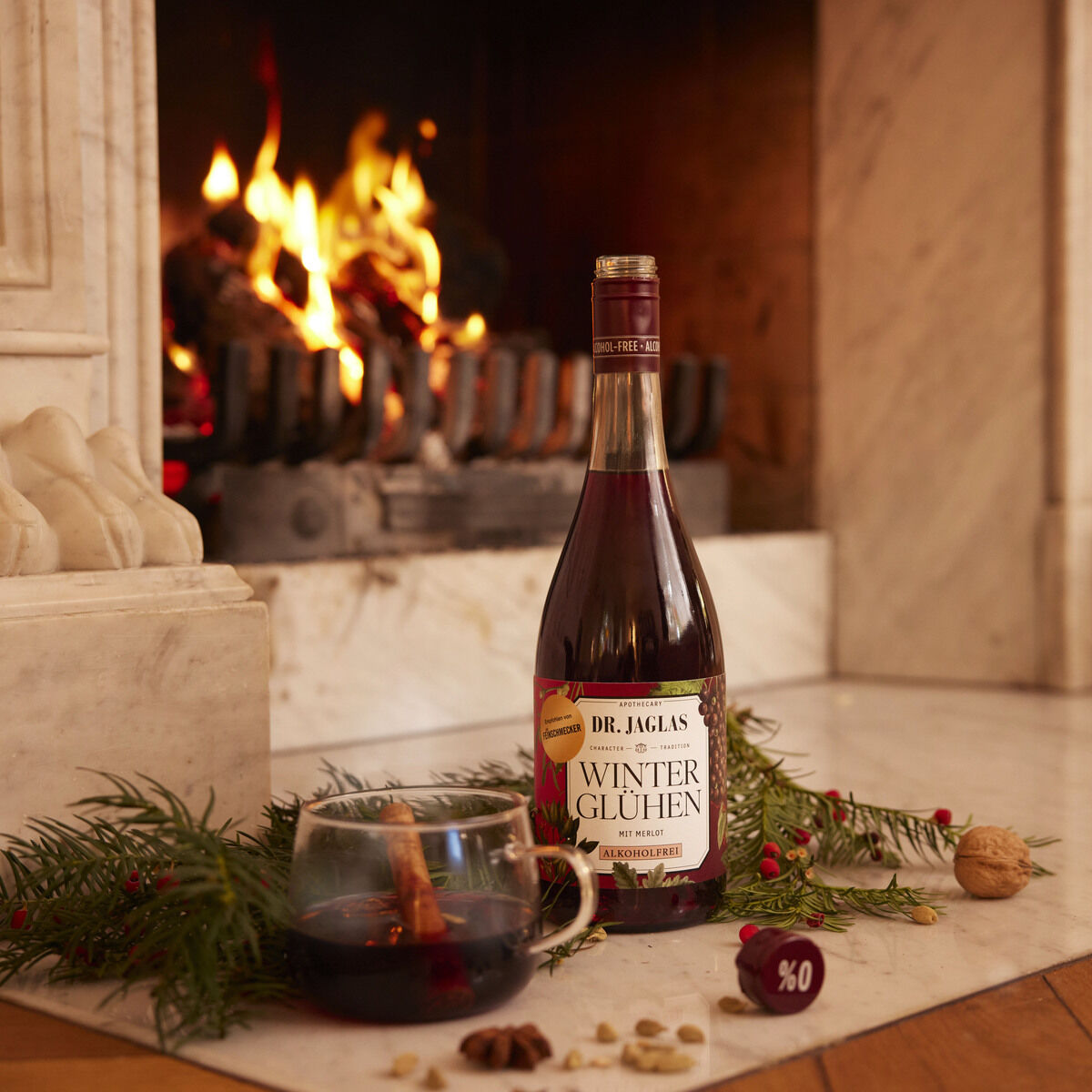 Flasche Winterglühen Merlot steht vor einem brennenden Kamin auf weißem Marmor. Daneben befinden sich ein Glas Rotwein, Tannenzweige und winterliche Dekoration wie Nüsse und Beeren. Im Hintergrund lodert das Feuer und verbreitet eine warme, gemütliche Stimmung.