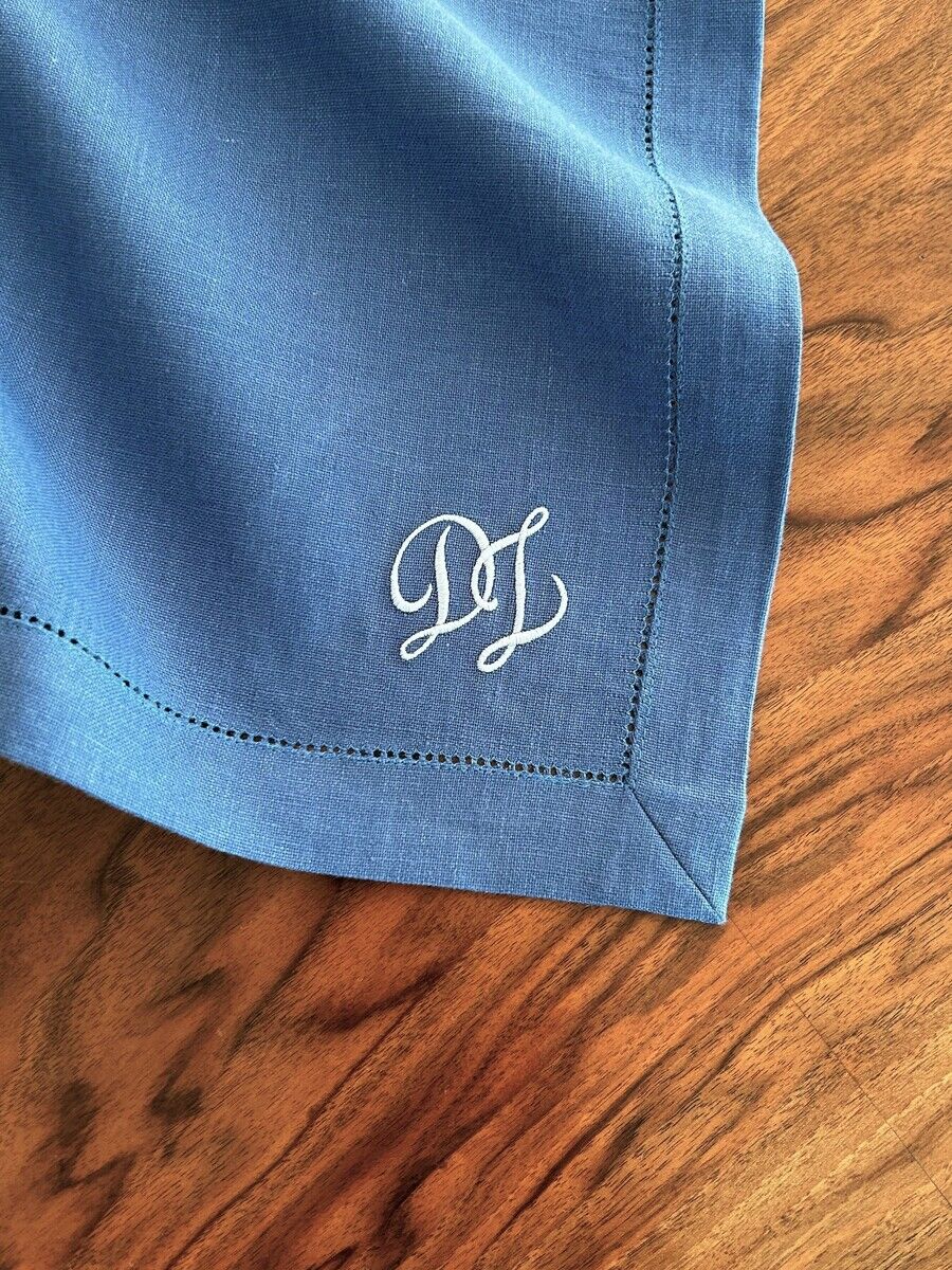 Eine blaue Leinenserviette mit einem weißen, handbestickten Monogramm auf einer Holztischplatte.