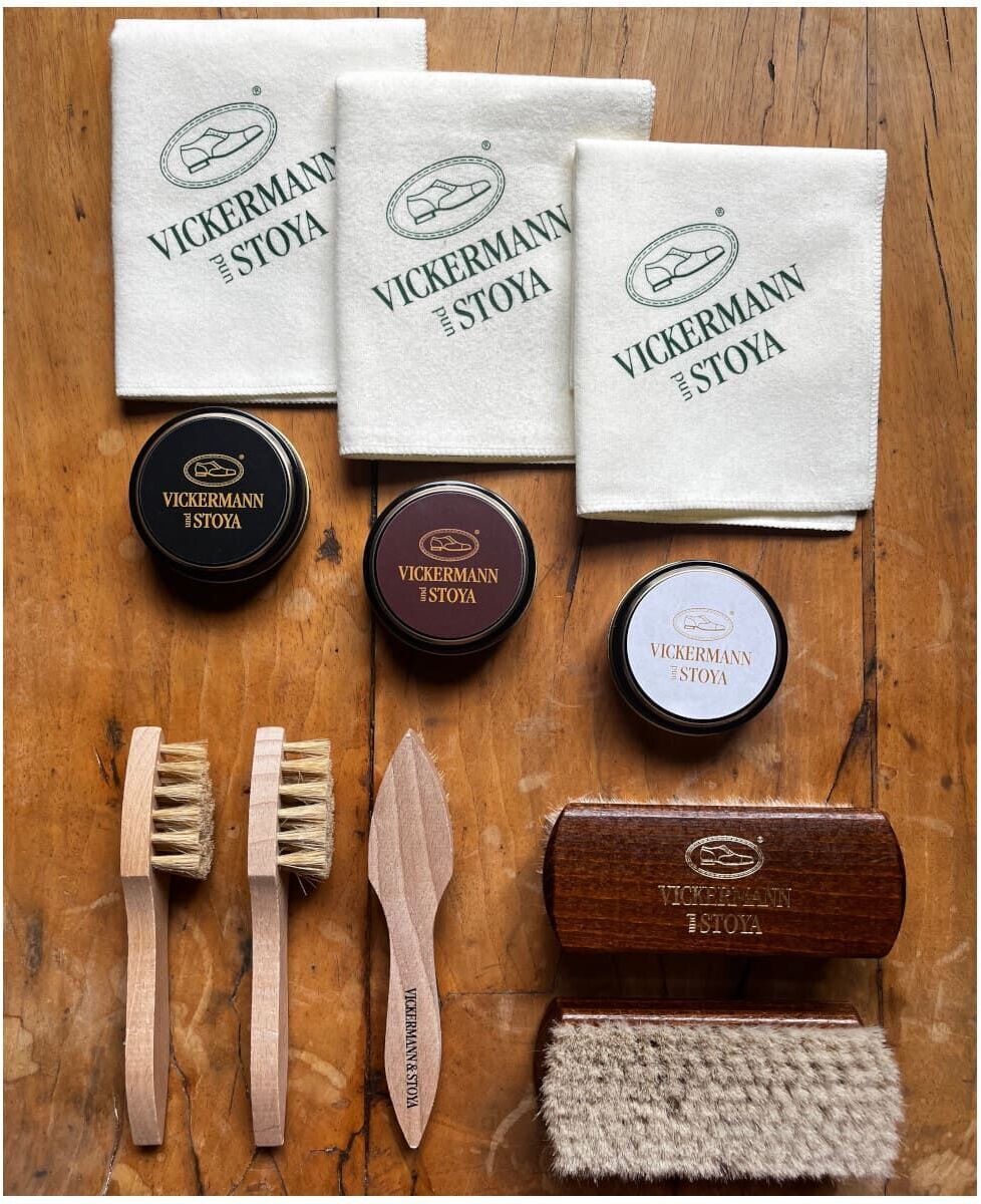 Lederpflege-Set von Vickermann & Stoya, bestehend aus Schuhcreme in drei Farben, vier Bürsten mit Holzgriffen, zwei Holzspateln und drei weißen Tüchern mit dem Vickermann & Stoya Logo, auf einer Holzoberfläche arrangiert.