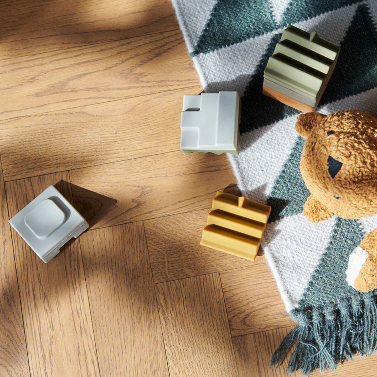 Mehrere farbige Holzblöcke des Stapelturms »Morgan« liegen auf einem Holzboden neben einem Stoffbären und einer gemusterten Decke.