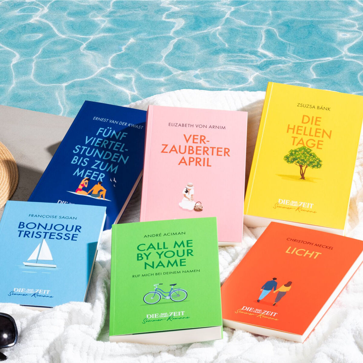 Eine Sammlung von sechs farbenfrohen Büchern der ZEIT-Edition »Sommer-Romane« liegt auf einem weißen Handtuch neben einem Swimmingpool. Die Bücher haben farbige Einbände in Blau, Gelb, Orange, Pink und Grün. Sonnenlicht reflektiert im Wasser des Pools, was eine entspannende Sommeratmosphäre schafft.