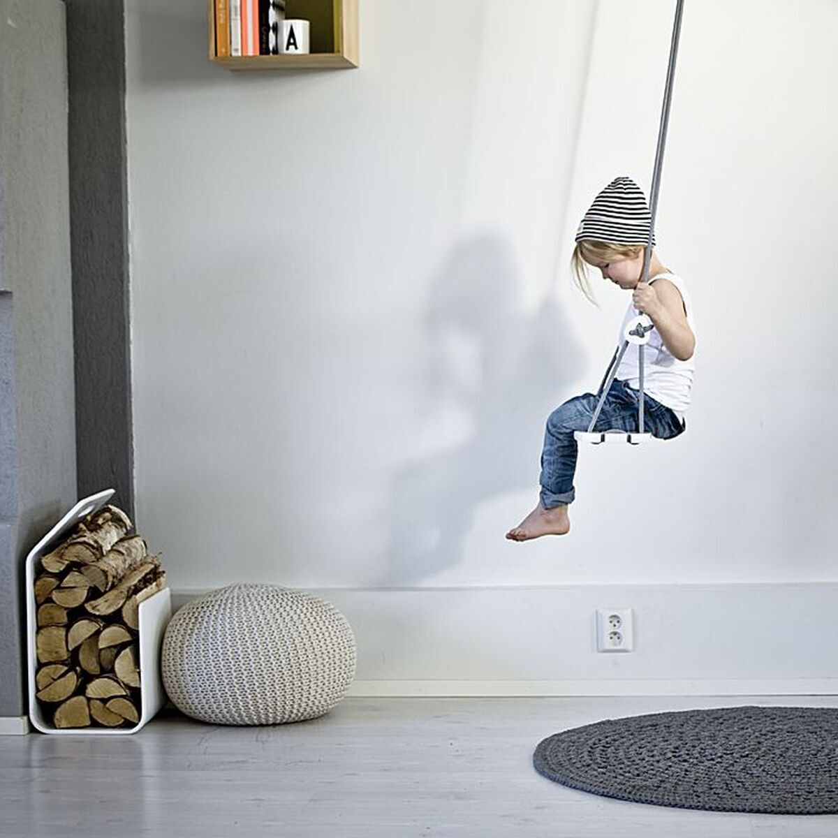 Ein Kind schaukelt auf einer modernen Kinderschaukel »Classic« im hell eingerichteten Wohnzimmer. Die Schaukel hängt an der Decke, im Raum befinden sich ein runder Teppich, ein Hocker und ein Holzstapel neben einer grauen Wand. Das Ambiente wirkt freundlich und minimalistisch.