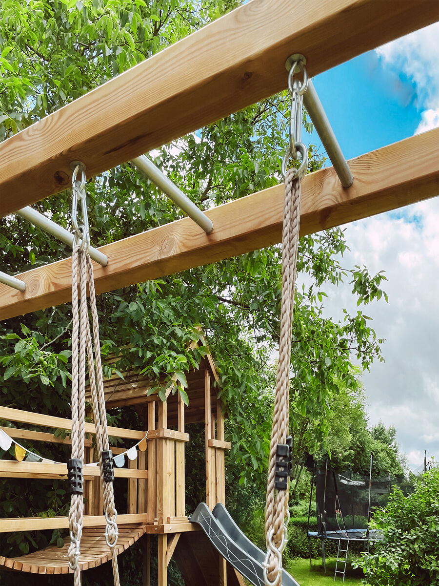 Ein Holz-Spielplatz mit einer Skateboard-Schaukel, Seilen und einer Rutsche im Garten bei sonnigem Wetter.